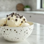 A scoop of smooth ice cream in a ceramic bowl, topped with chocolate chips and placed on a marble countertop in a modern kitchen.